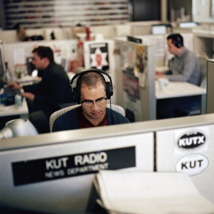 KUT radio staff member wearing headphones at workstation in open-plan newsroom.