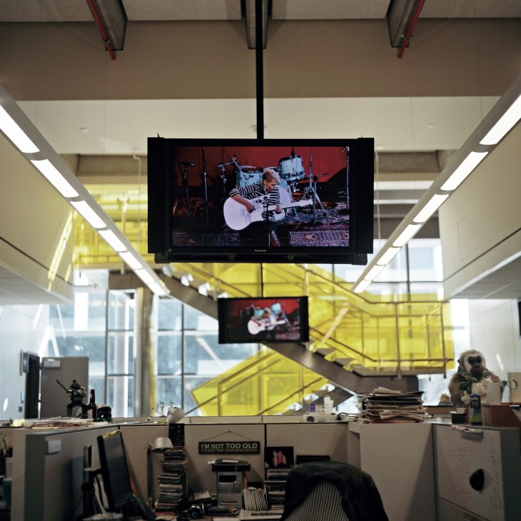 TV screens suspended from ceiling display live musical performances in a radio station studio