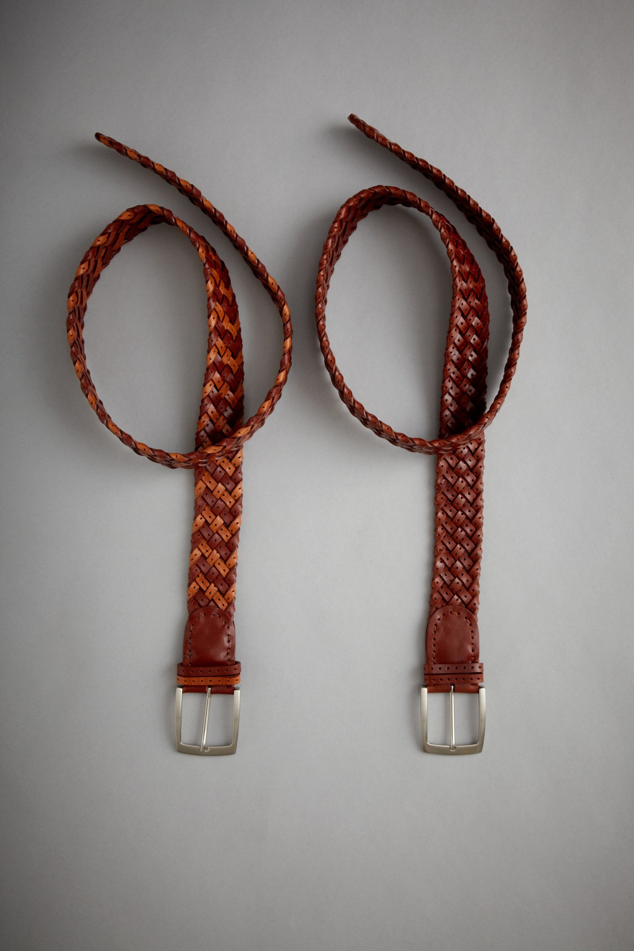 Two woven leather belts in cognac brown with metal buckles displayed against a grey background.
