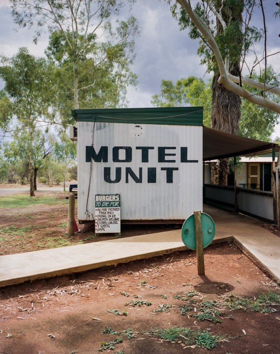 A roadside motel unit with green awning advertising burgers, set among eucalyptus trees in the Australian outback.