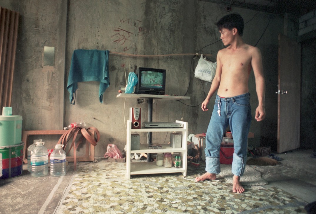 A shirtless Burmese migrant stands in a sparse, deteriorating concrete room with a television on a makeshift shelf.