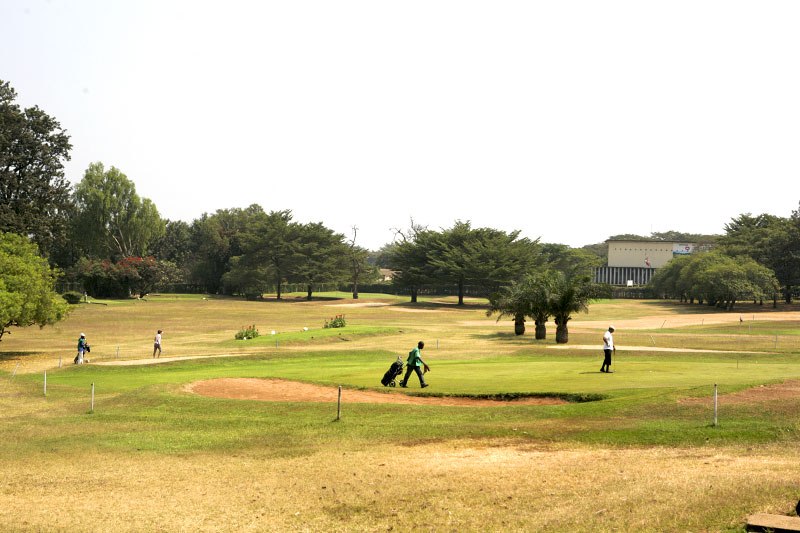 Players on a well-maintained golf course with trees and clubhouse in background.