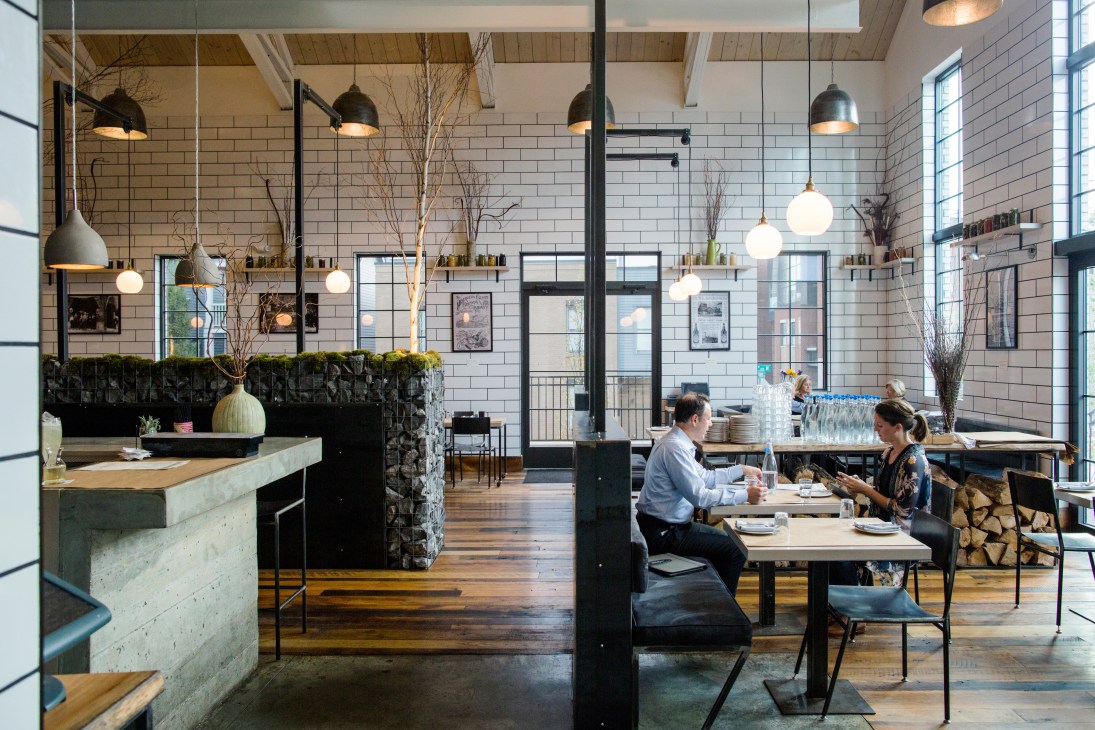 Industrial-style restaurant interior with white subway tiles, dark pendant lights, and diners seated at wooden tables.