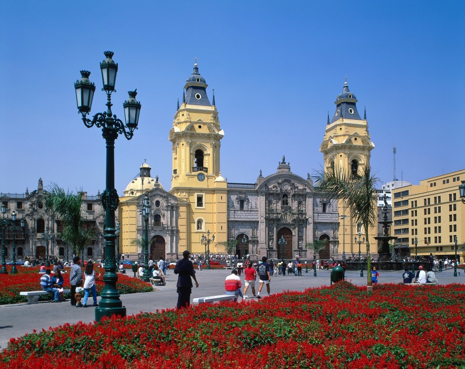 Lima's central plaza with yellow colonial churches, ornate street lamp, and red flowers under clear blue sky.