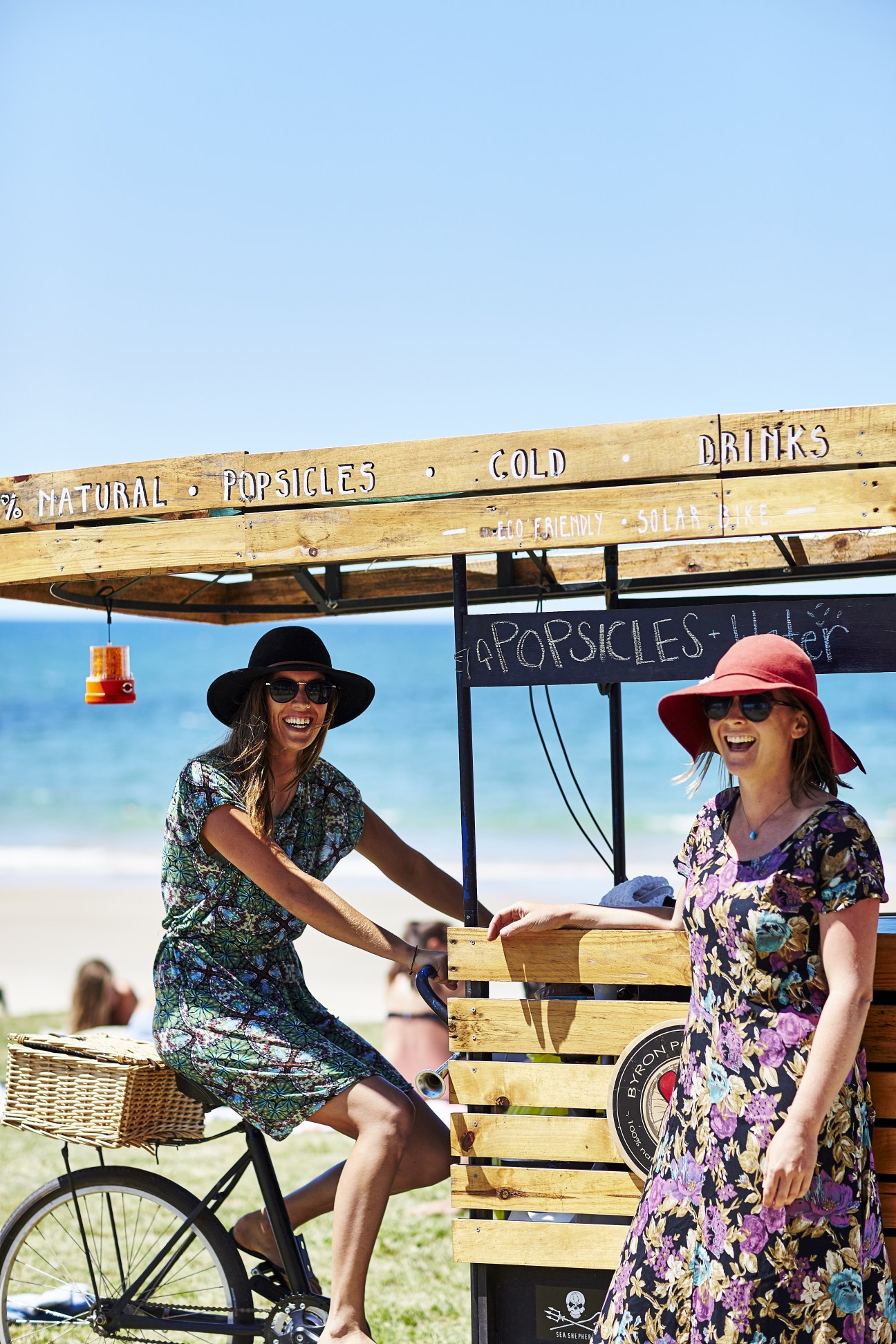 Two women at a wooden popsicle stand on a beach with ocean in the background