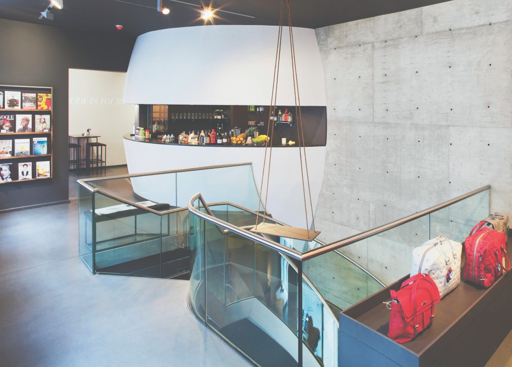 Modern retail interior with concrete walls, glass railings, suspended white counter with bar seating, and display cases with red backpacks.