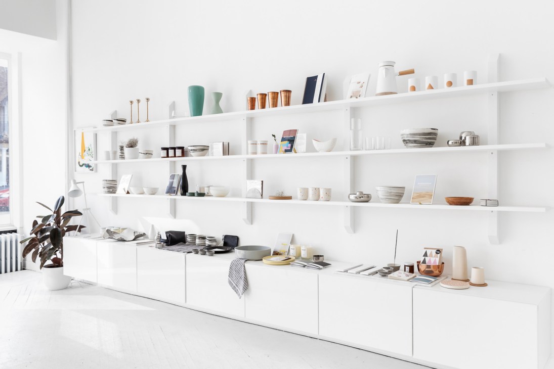 White shelving unit displaying various ceramic vessels, books, and design objects in a minimalist retail setting