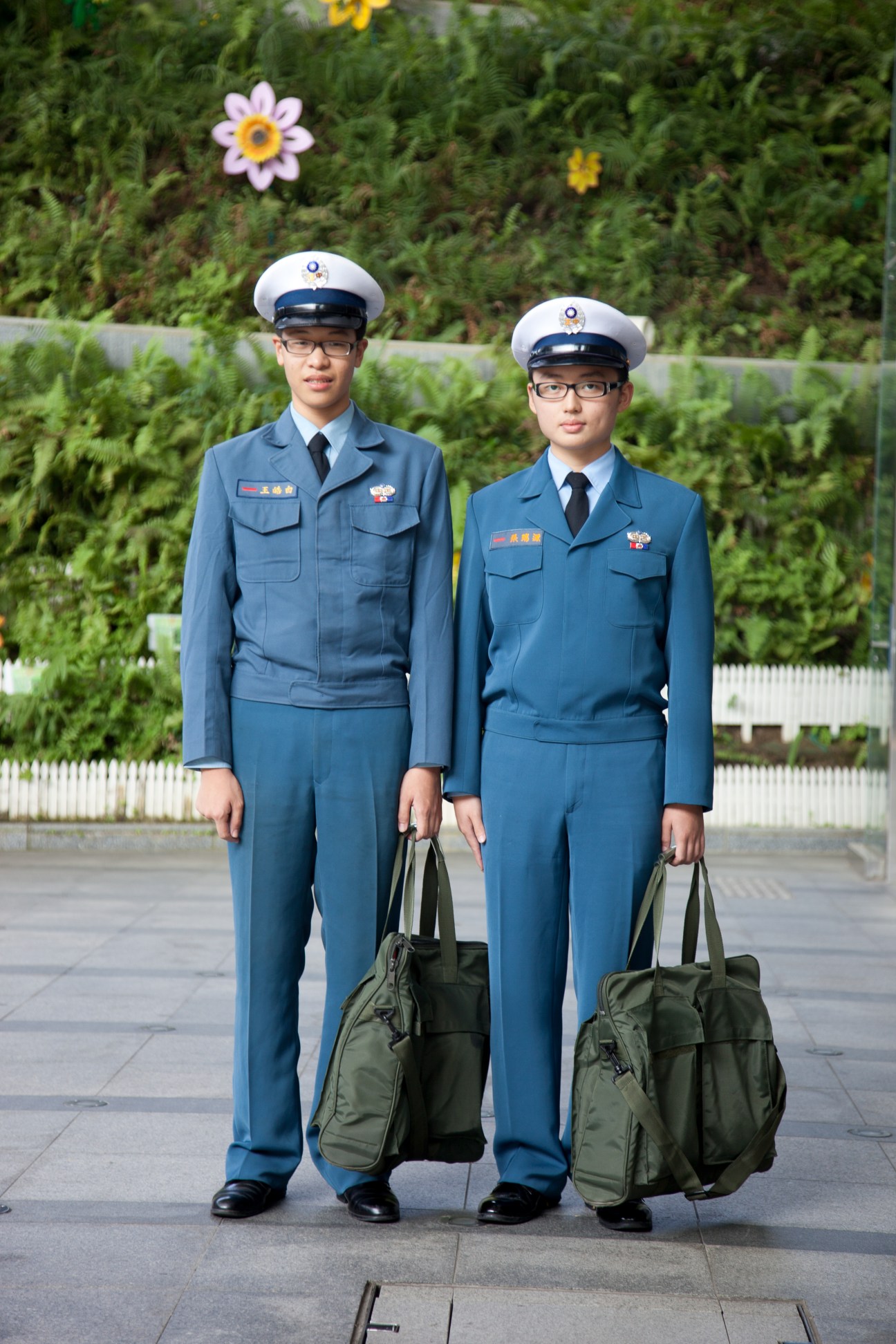Two young military cadets in blue uniforms and white caps stand holding olive green bags.