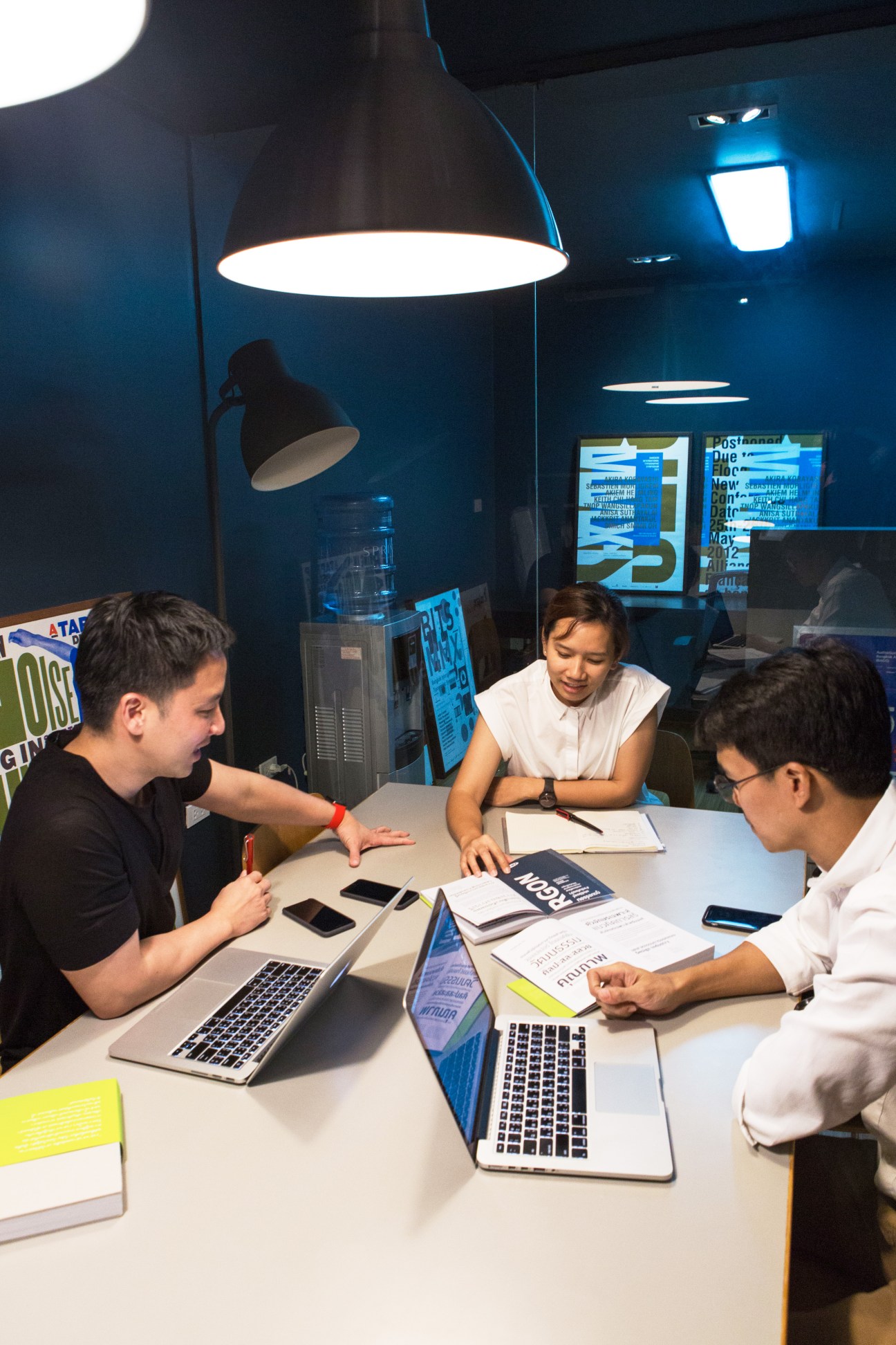 Three people collaborating at a modern office table with laptops, documents, and design materials under pendant lighting
