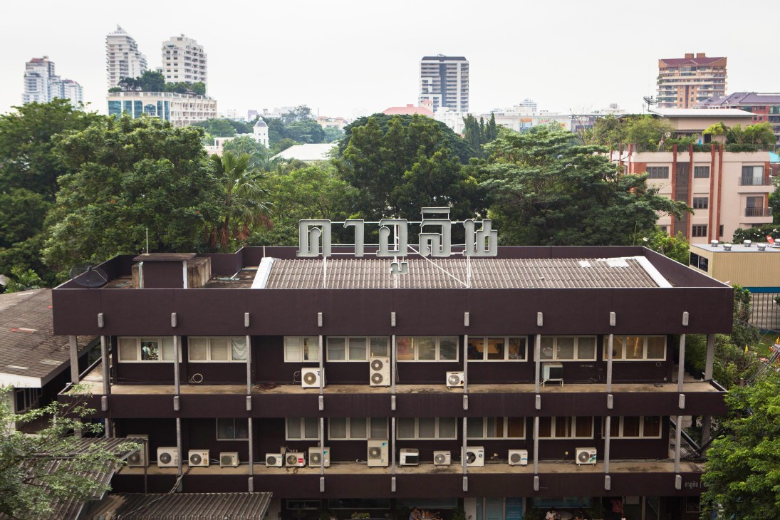 Modern brutalist-style concrete building with multiple air conditioning units and rooftop signage in urban Bangkok