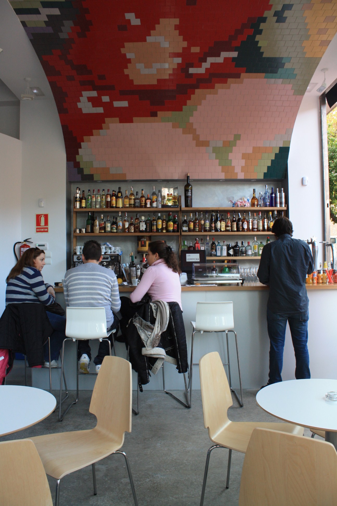 Interior of Café Lempicka showing bar with bottles and pixelated ceiling mosaic art in red and earth tones.