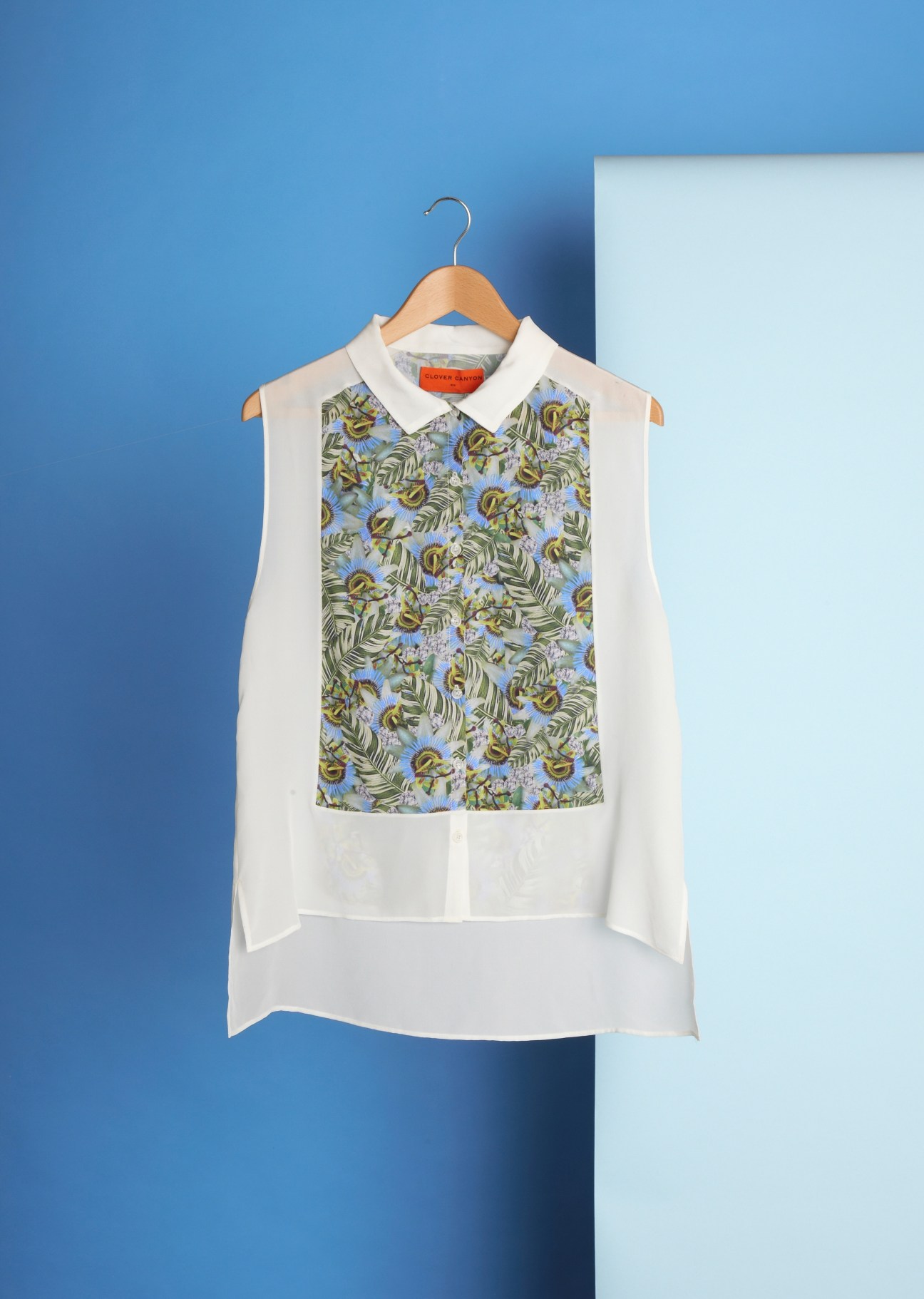 Sleeveless white shirt with floral print panel on wooden hanger against blue background.
