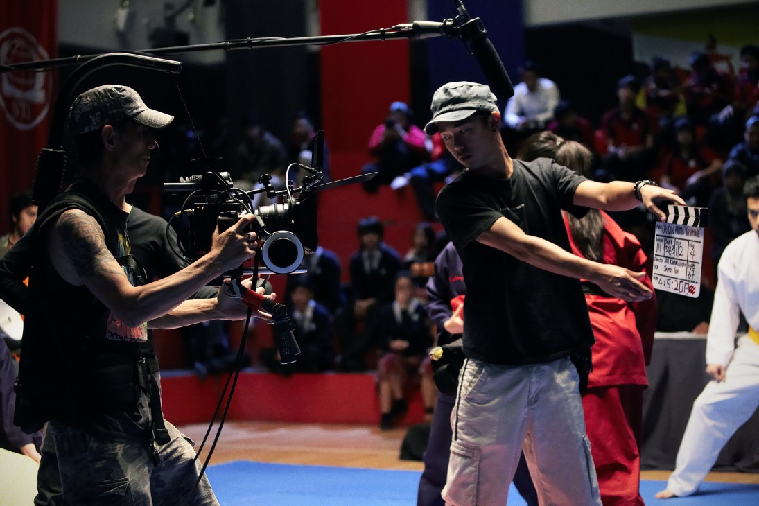 Camera crew operating professional film equipment on set during a university auditorium scene with extras and crowd in background.