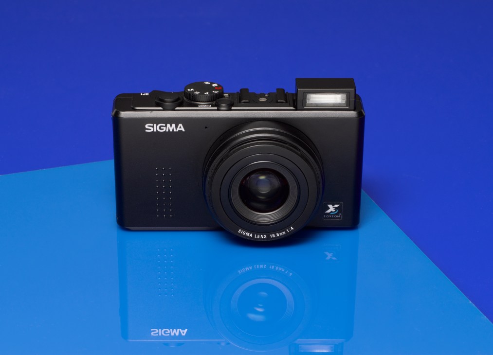 Sigma DP-1 compact digital camera with black body and lens on blue gradient background.