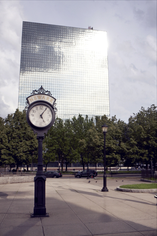 A vintage street clock in Newark's Military Park with a modern glass skyscraper and trees in the background.