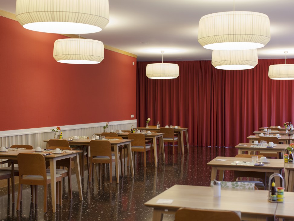 Neugut care home dining hall with wooden tables, chairs, pendant lights, red walls and burgundy curtains.