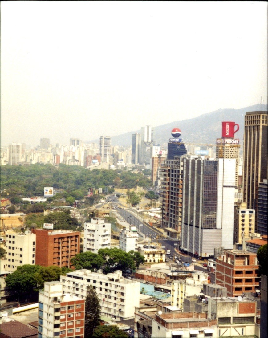 Aerial view of downtown Caracas with high-rise buildings, mountains in background, and lush green vegetation in the valley.