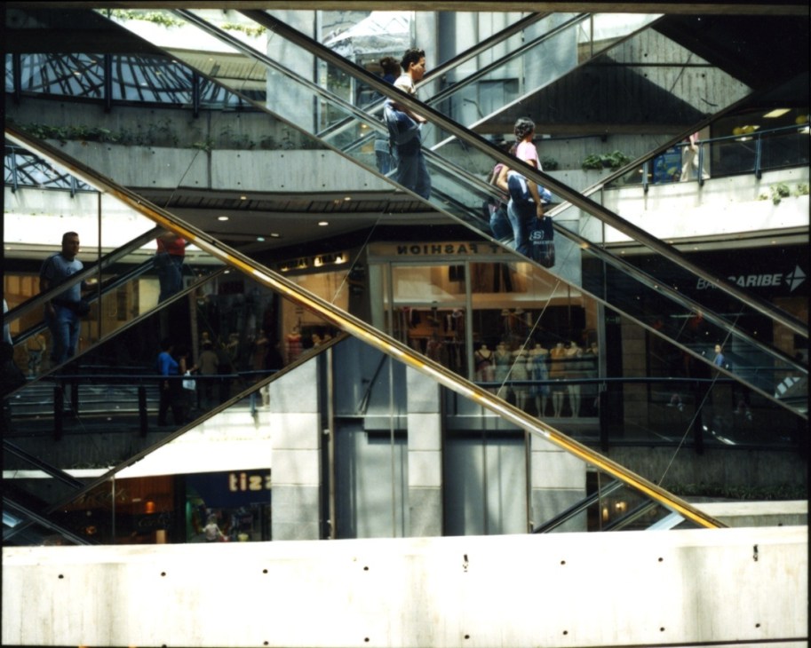 Shoppers on escalators inside a modern Caracas shopping mall with concrete architecture.
