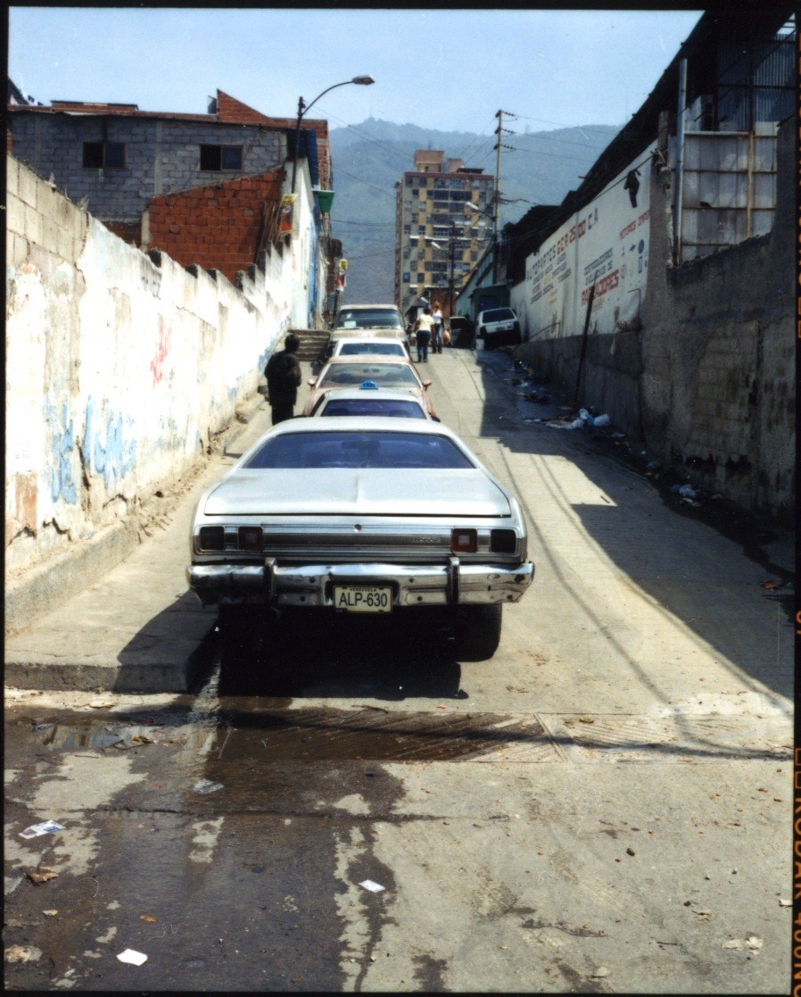 Line of white cars parked on a narrow street in Caracas with mountains visible in the distance.