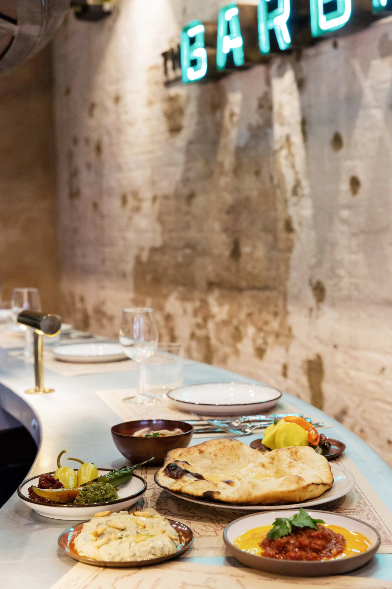 The Barbary restaurant dining table with naan bread, mezze dishes, and turquoise neon sign on stone wall.