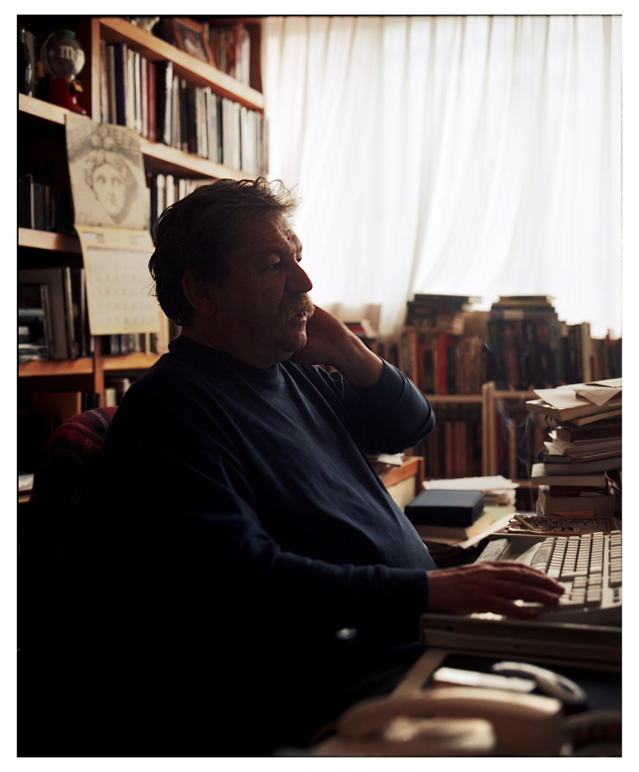 Paco Ignacio Taibo II sits at his desk with a keyboard, looking thoughtfully to the side in his study lined with bookshelves.