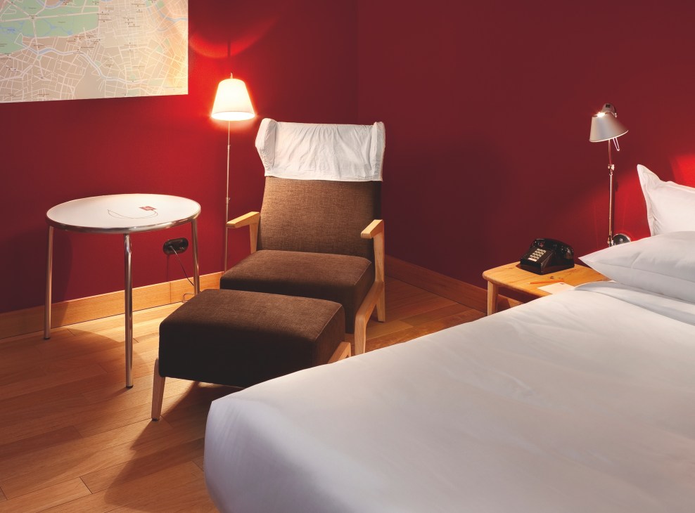 Modern hotel room with bold red walls, wooden furniture, white bedding, and contemporary lighting fixtures.