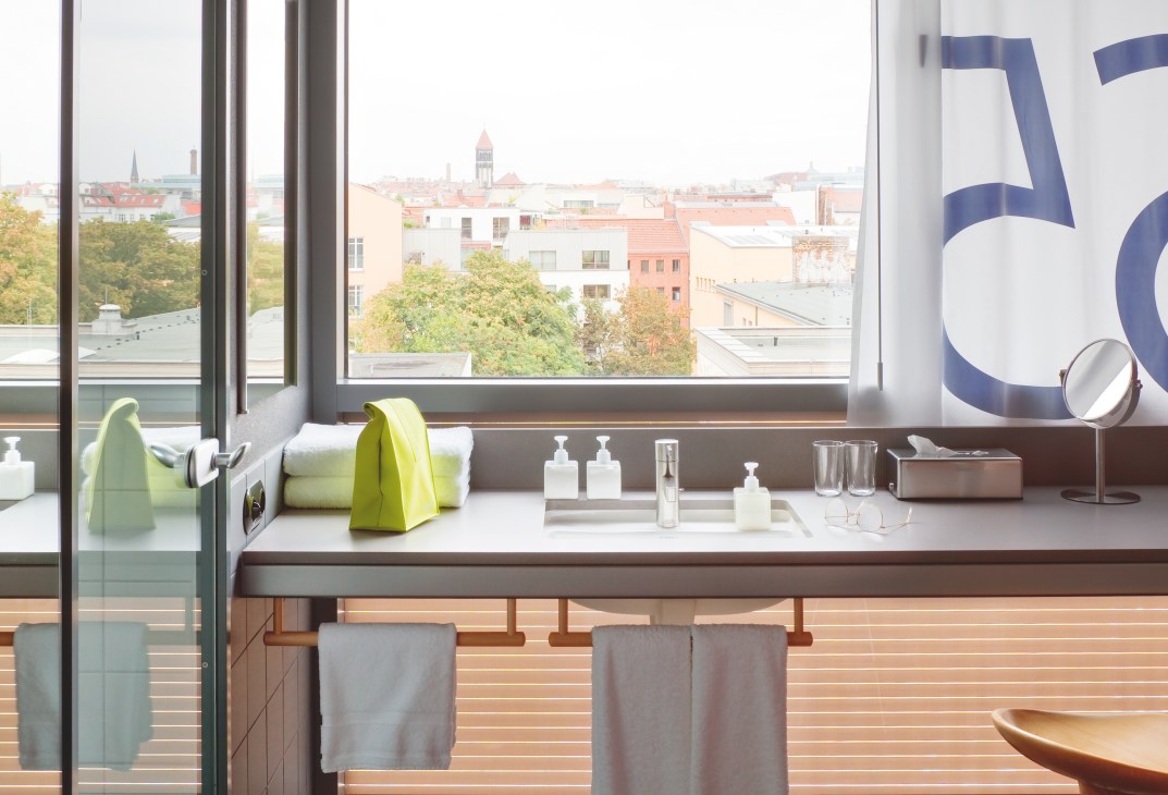 Modern hotel bathroom vanity with city skyline view through large windows, featuring lime green accessories and cosmetic products.