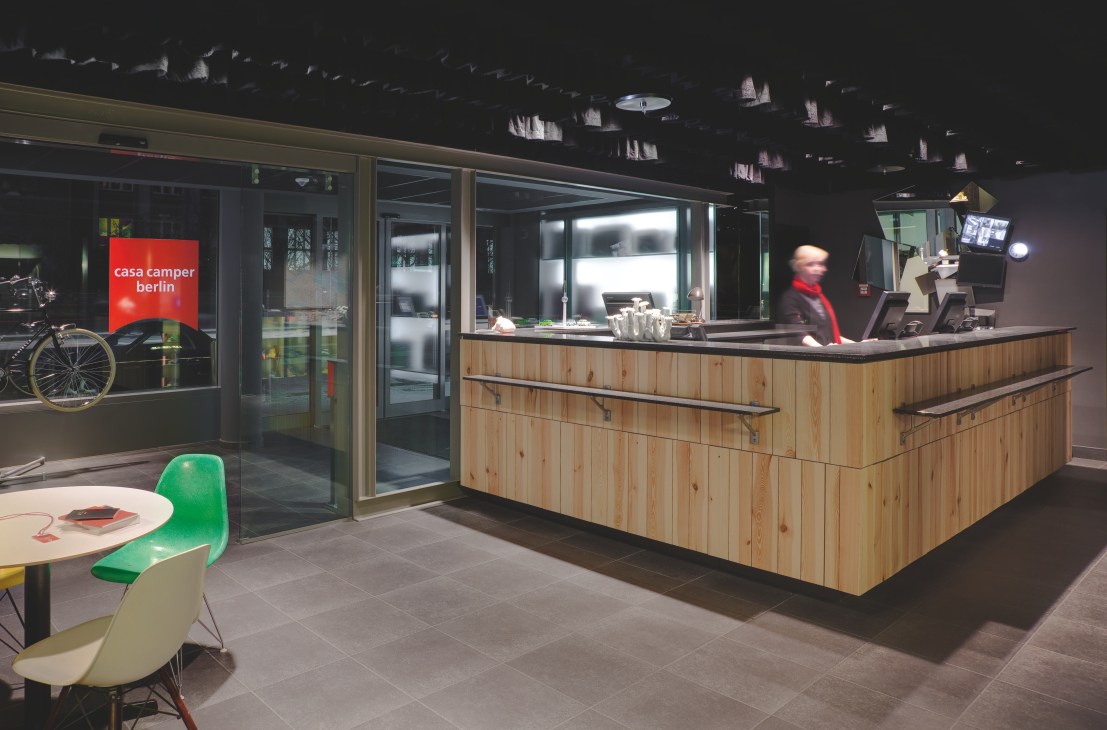 CasaCamper Berlin reception desk with wooden paneling, staff member, and green chair in modern hotel lobby.