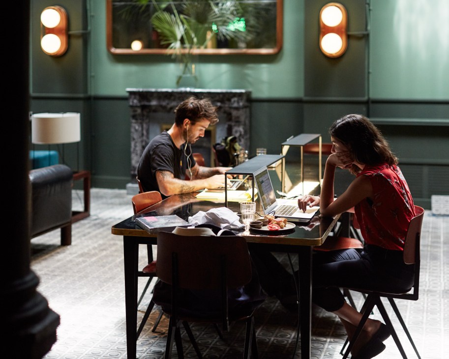 Two people work at laptops at a table in a modern hotel lobby with green walls and warm lighting.