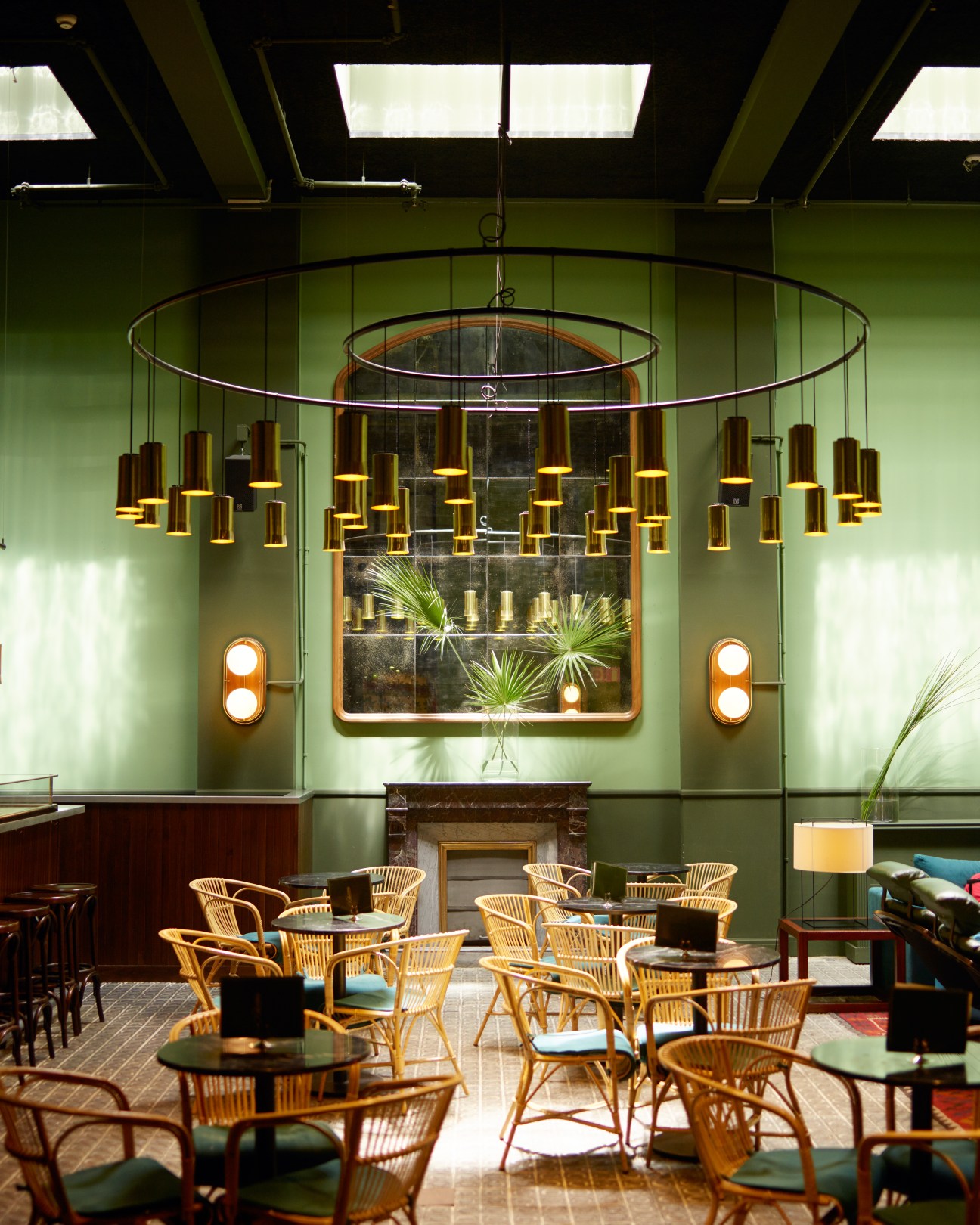 A modernist lounge with sage green walls, rattan chairs, and a dramatic brass pendant light fixture.