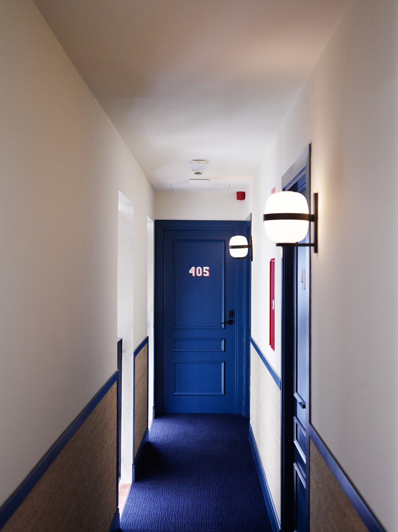 Hotel corridor with blue door numbered 405, white walls, blue carpet, and modern wall sconces.