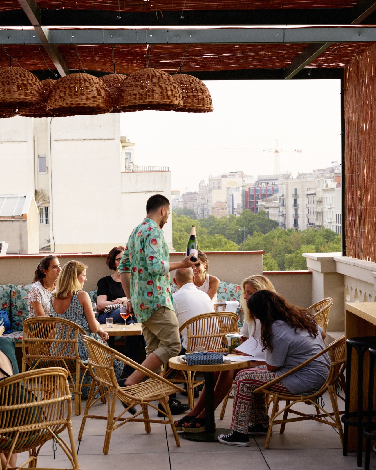 People socializing on a rooftop terrace at Casa Bonay with Barcelona cityscape views and wicker pendant lights overhead.