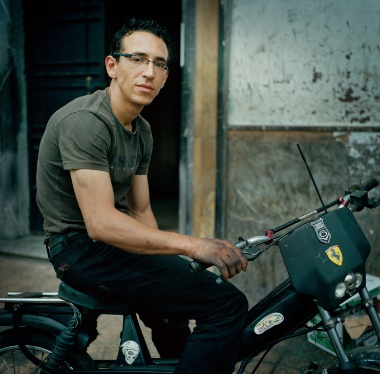 Man sitting on a motorcycle in a workshop with weathered walls behind him.