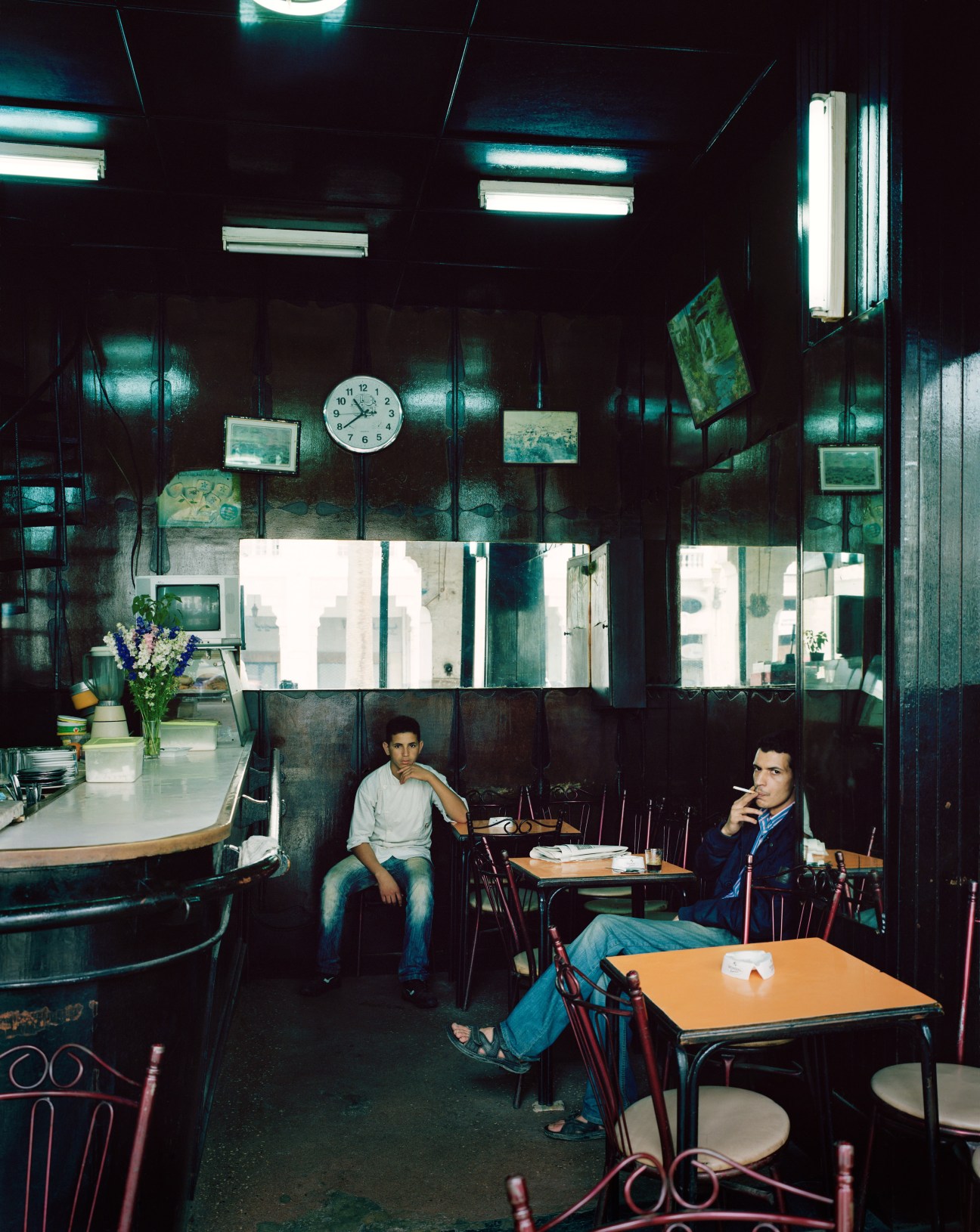 Two men sit in a dimly lit café with fluorescent lighting, large windows, and a clock on the wall.