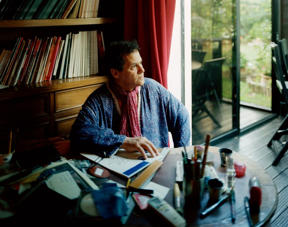 Mehdi Qotbi sits at his desk by a window, surrounded by bookshelves and art supplies.