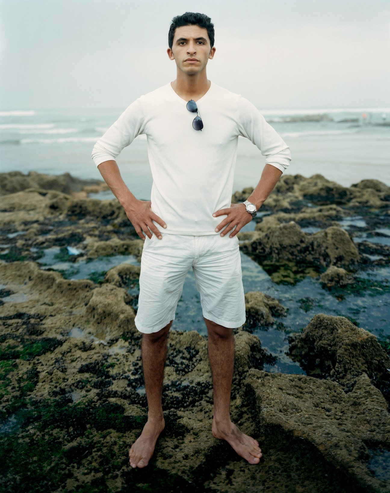 Man standing on rocky beach wearing white shirt and shorts, sunglasses around neck, ocean behind him.