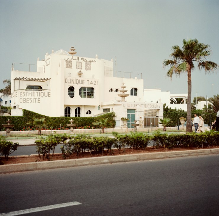 White Art Deco building with 'Clinique Tazi' signage, palm trees, and manicured hedges on Boulevard de la Corniche.