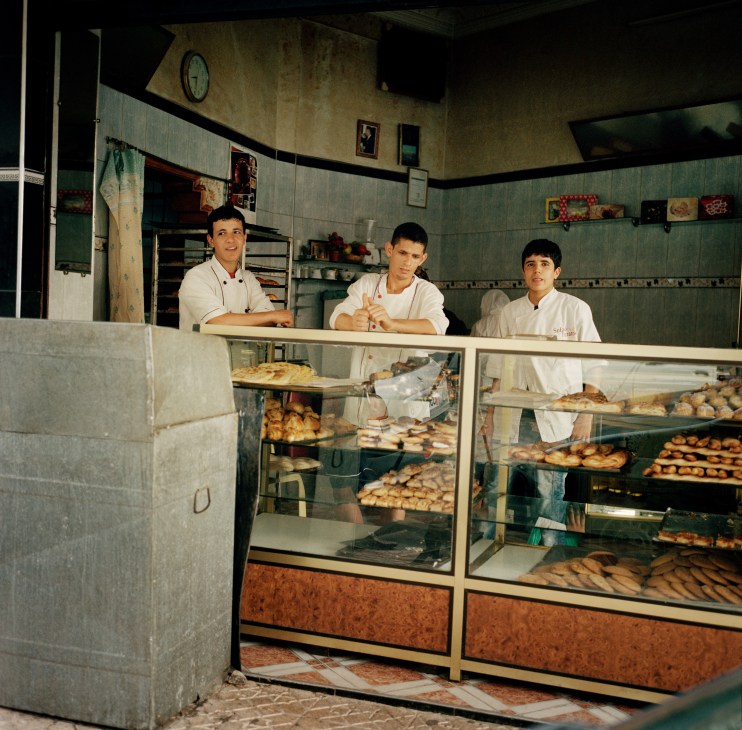 Three bakers in white uniforms stand behind a counter in a modest bakery with tiled walls and display cases of fresh bread.
