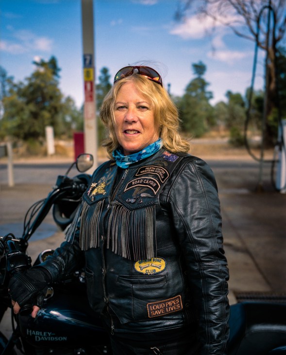 Woman in black leather jacket and sunglasses stands beside a Harley-Davidson motorcycle at a roadhouse.