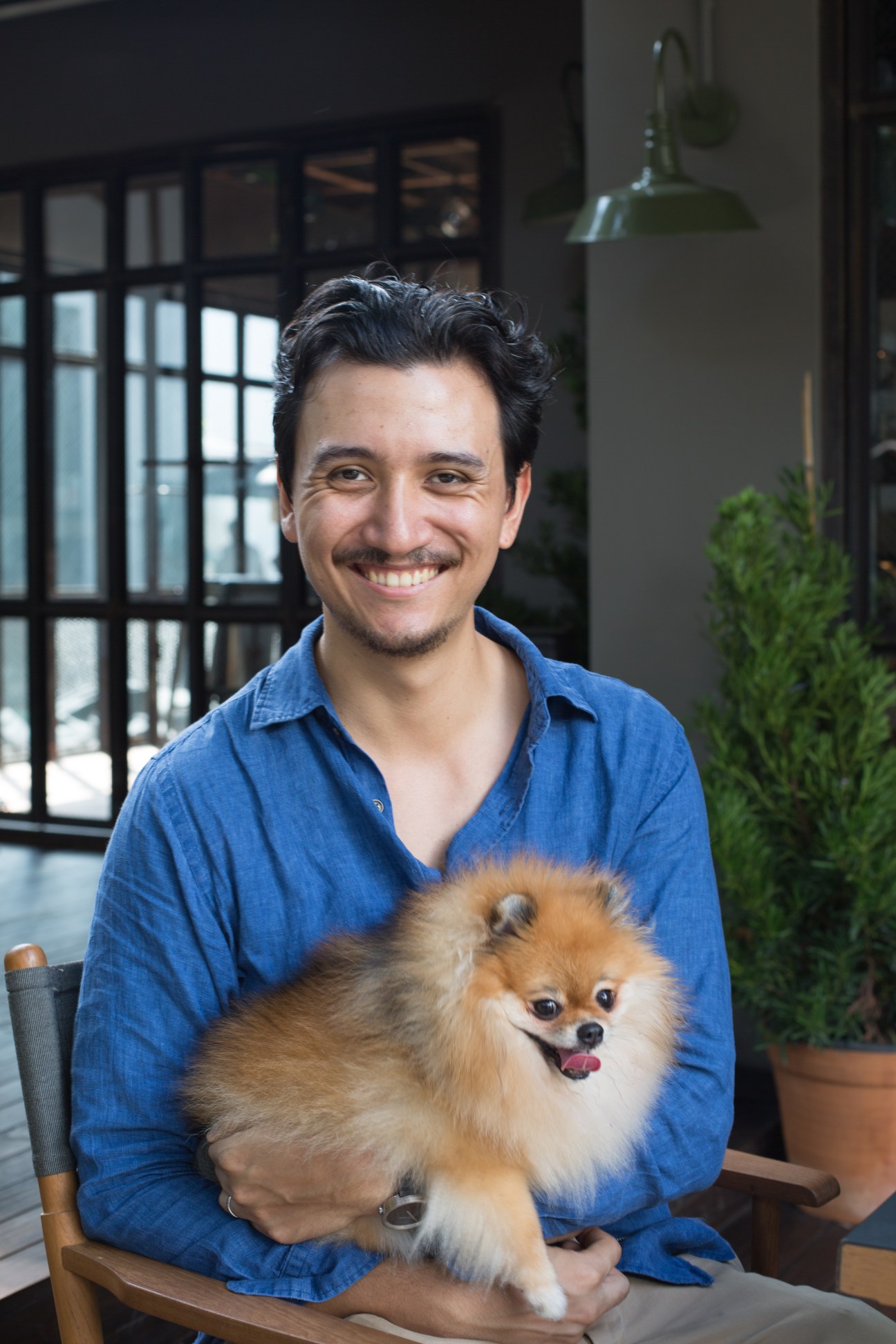 Man in blue shirt holding a fluffy tan and white Pomeranian dog while sitting outdoors.