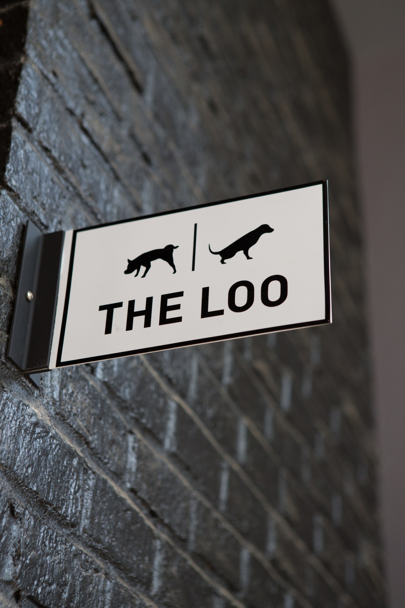 Sign reading 'The Loo' with dog silhouettes, mounted on a textured concrete wall.