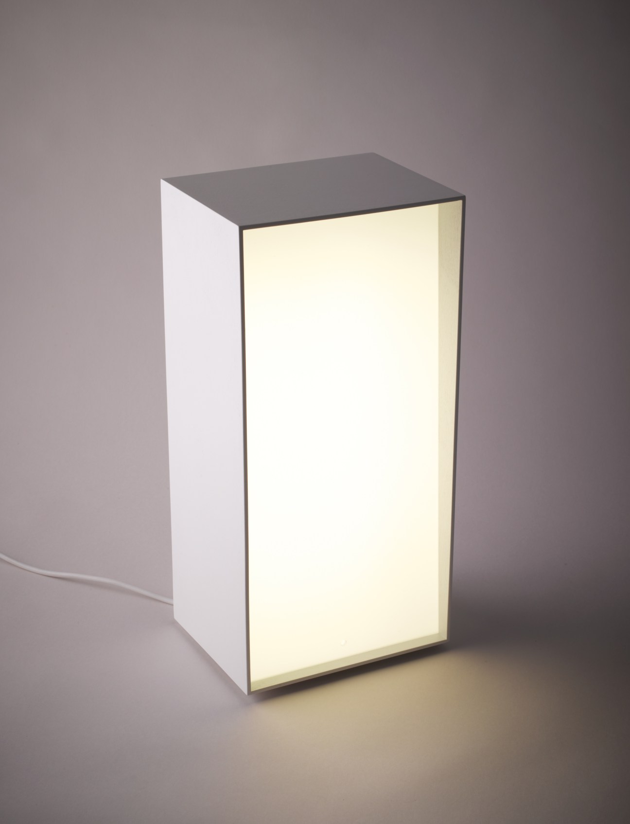 A minimalist rectangular table lamp with a dark top and illuminated cream-colored side panel.