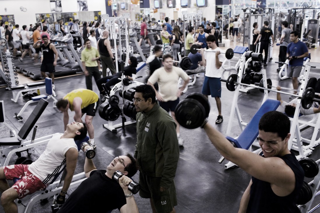 Busy gymnasium with multiple people exercising with dumbbells and using various fitness equipment.