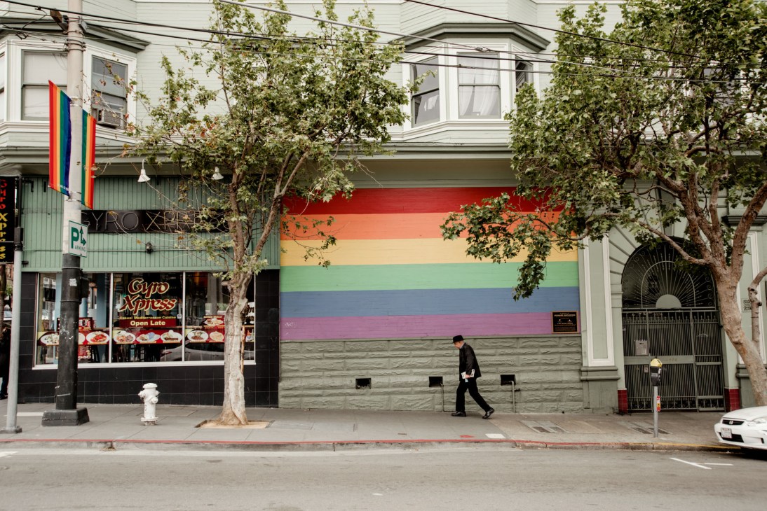 A person walks past a San Francisco storefront with a large rainbow pride flag mural and a cafe sign.