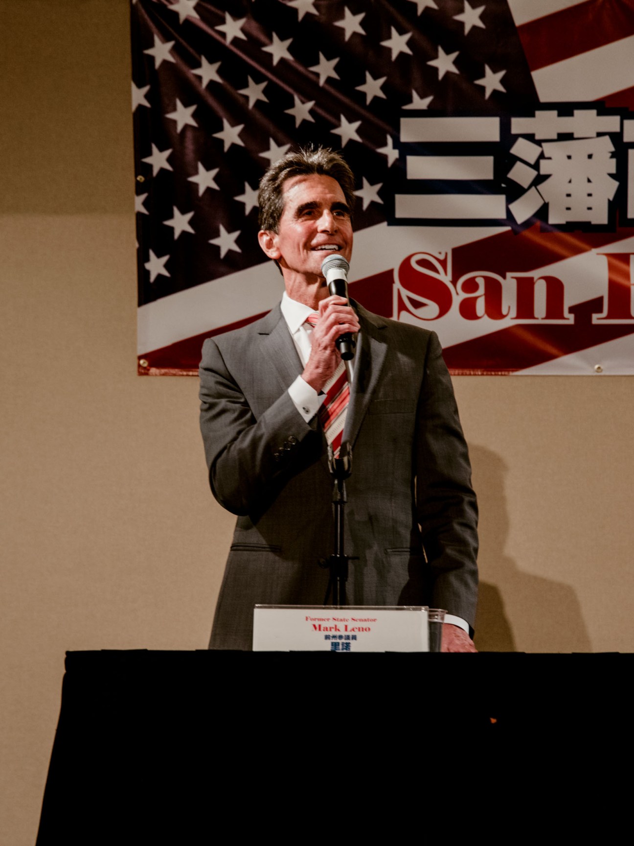 Former state senator Mark Leno speaking at a podium with American flag backdrop.