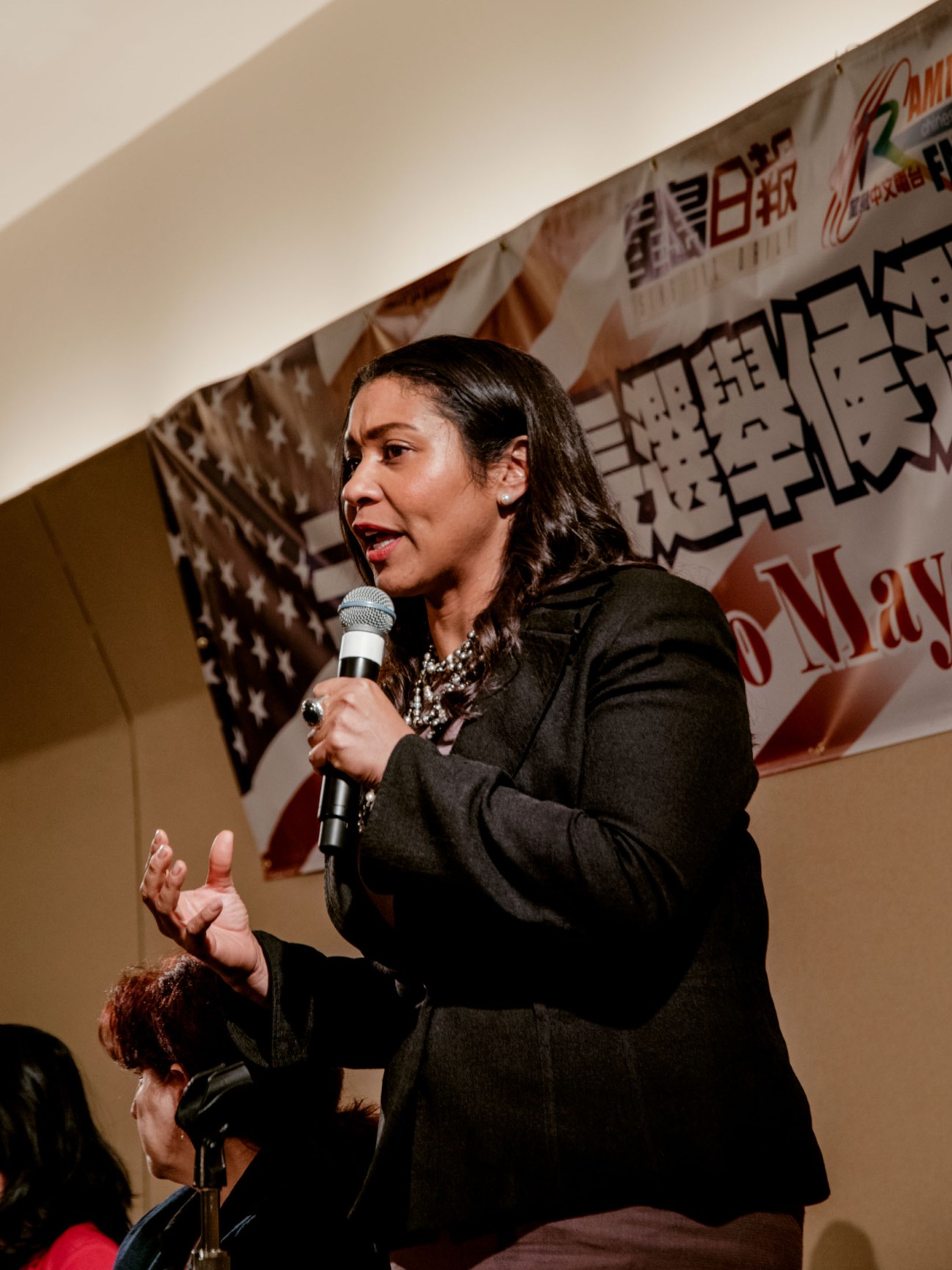 London Breed speaking at a microphone during a campaign event with banners behind her.