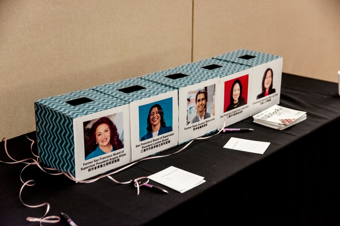 A decorative voting booth display featuring portraits of four San Francisco mayoral candidates with bilingual labels on a black table.