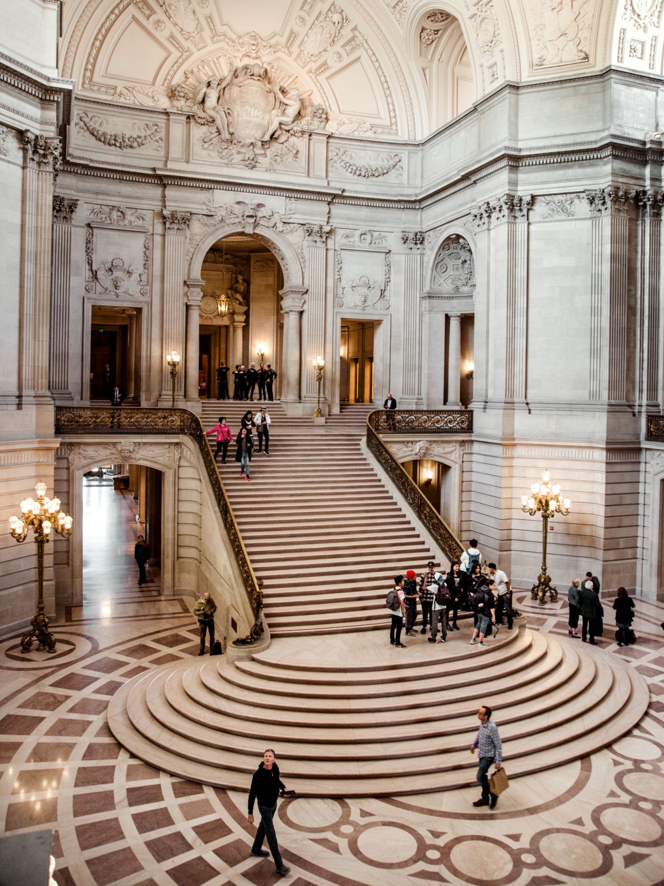 Interior rotunda and grand staircase of San Francisco City Hall with ornate neoclassical architecture and visitors.