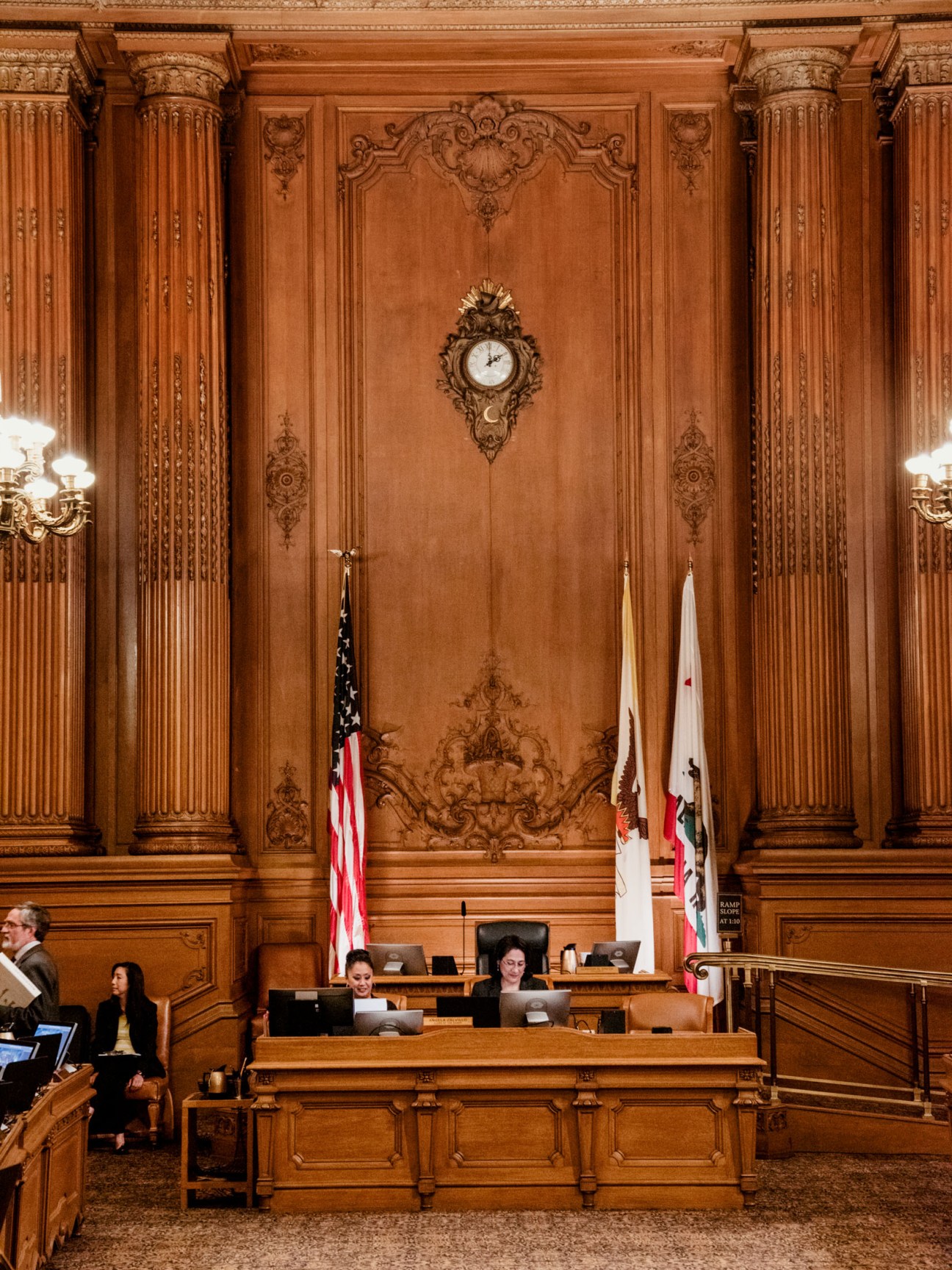 Interior of San Francisco City Hall courtroom with ornate wooden paneling, columns, clock, and American and California flags.