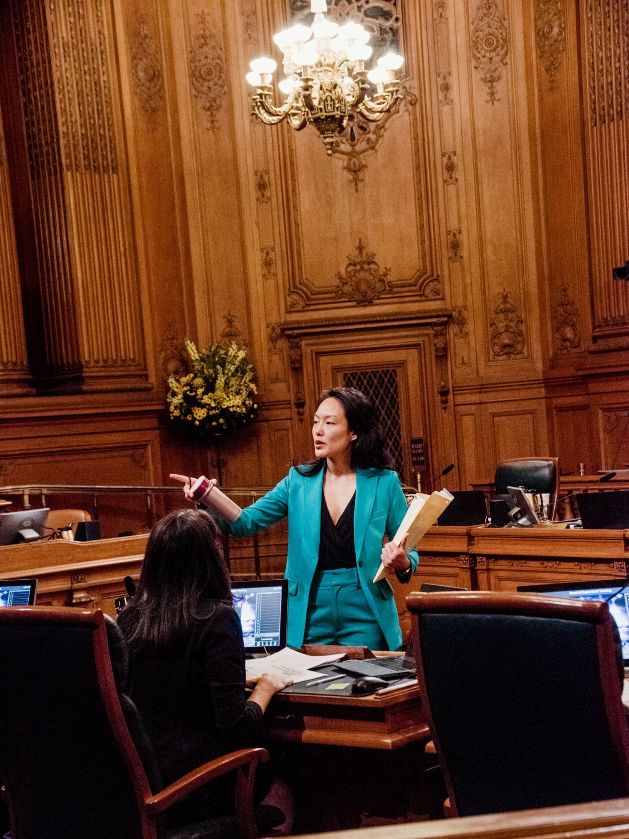 Woman in turquoise blazer gesturing while speaking in an ornate wooden courtroom.